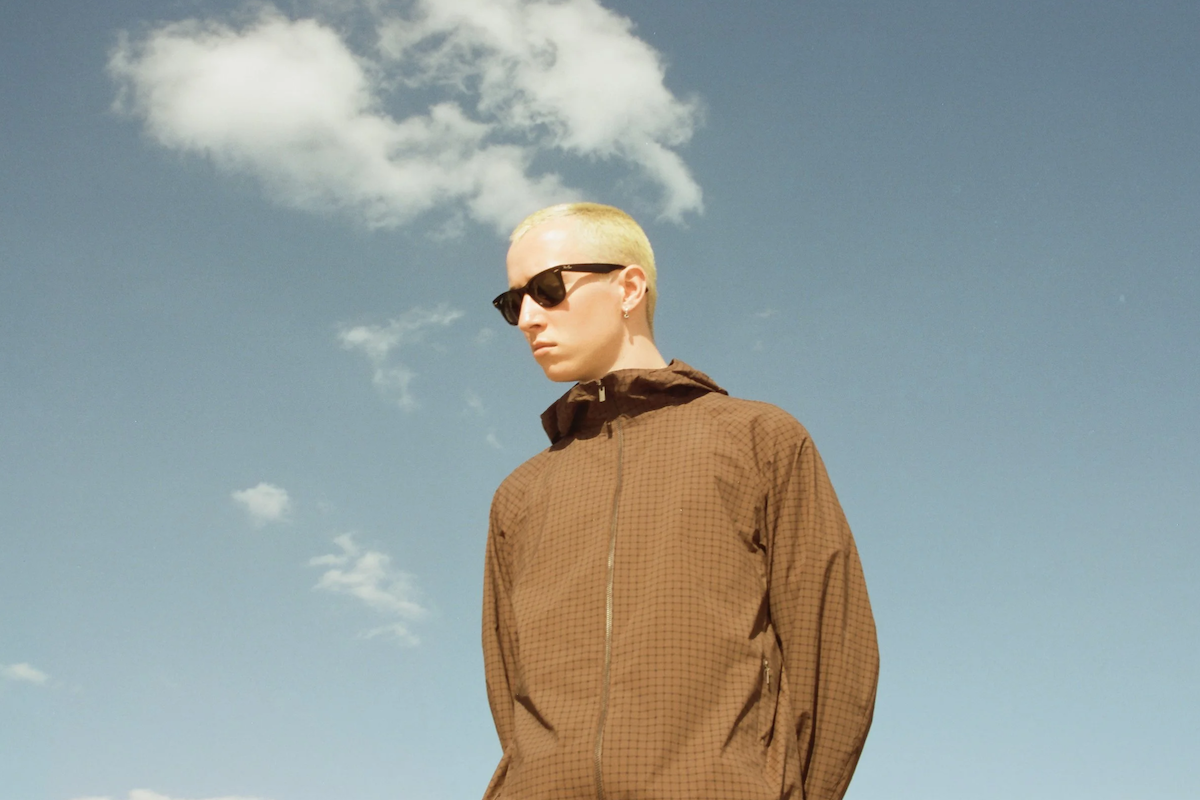 Musician Jack Kaye wearing sunglasses and a brown jacket, posing outdoors against a blue sky with clouds.